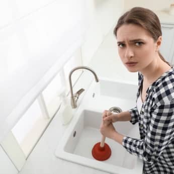 upset-young-woman-using-plunger-unclog-sink-drain-kitchen-view